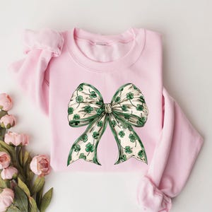 May include: A light pink sweatshirt featuring a large bow design. The bow has a white background with green shamrocks and a green border. The sweatshirt is a crew neck style with long sleeves.