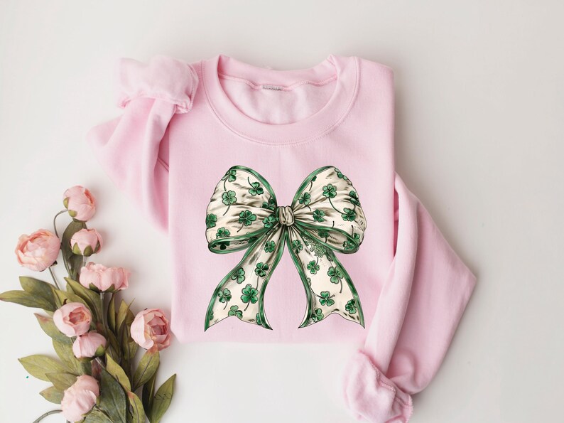 May include: A light pink sweatshirt featuring a large bow design. The bow has a white background with green shamrocks and a green border. The sweatshirt is a crew neck style with long sleeves.