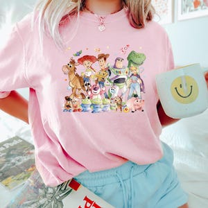 May include: A light pink t-shirt featuring a colorful graphic of characters from the animated movie Toy Story. The design includes Woody, Buzz Lightyear, Jessie, and other beloved characters. The shirt is paired with light blue shorts.