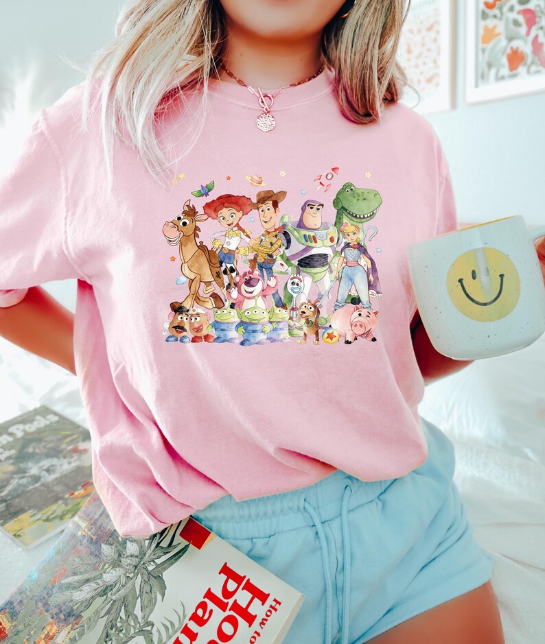 May include: A light pink t-shirt featuring a colorful graphic of characters from the animated movie Toy Story. The design includes Woody, Buzz Lightyear, Jessie, and other beloved characters. The shirt is paired with light blue shorts.