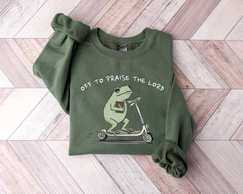 May include: An olive green sweatshirt with the phrase "Off to Praise the Lord" printed above a cartoon frog riding a scooter and holding a book. The sweatshirt has long sleeves and a crew neck.