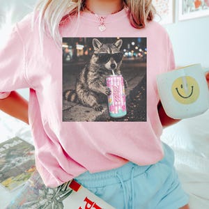 May include: A light pink sweatshirt with a graphic of a raccoon wearing sunglasses and drinking from a can. The can has the word "Always" on it. The person is also holding a mug with a yellow smiley face.