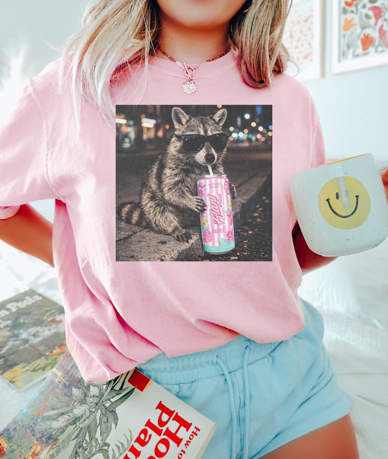 May include: A light pink sweatshirt with a graphic of a raccoon wearing sunglasses and drinking from a can. The can has the word "Always" on it. The person is also holding a mug with a yellow smiley face.
