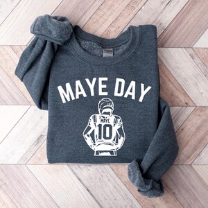 K&ouml;nnte beinhalten: Ein dunkelgraues Sweatshirt mit dem wei&szlig;en Schriftzug "MAYE DAY". Darunter befindet sich eine wei&szlig;e Grafik eines Footballspielers mit der Nummer 10. Das Sweatshirt liegt auf einer Holzoberfl&auml;che.