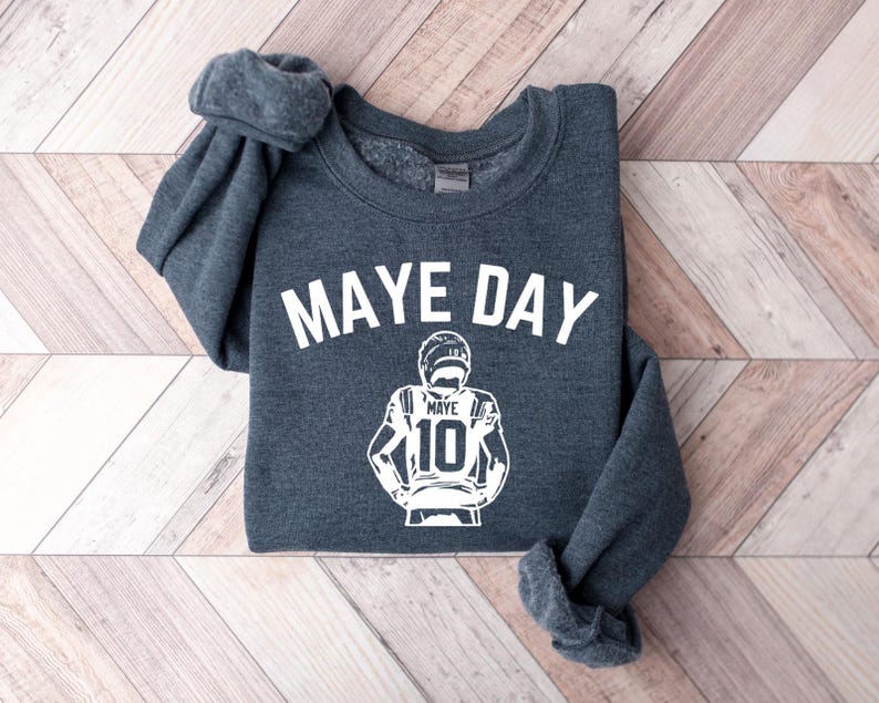 K&ouml;nnte beinhalten: Ein dunkelgraues Sweatshirt mit dem wei&szlig;en Schriftzug "MAYE DAY". Darunter befindet sich eine wei&szlig;e Grafik eines Footballspielers mit der Nummer 10. Das Sweatshirt liegt auf einer Holzoberfl&auml;che.