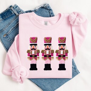 May include: A light pink sweatshirt featuring three embroidered nutcracker figures. The nutcrackers have pink and gold uniforms, black boots, and detailed facial features. The sweatshirt is laid on top of a pair of blue jeans.