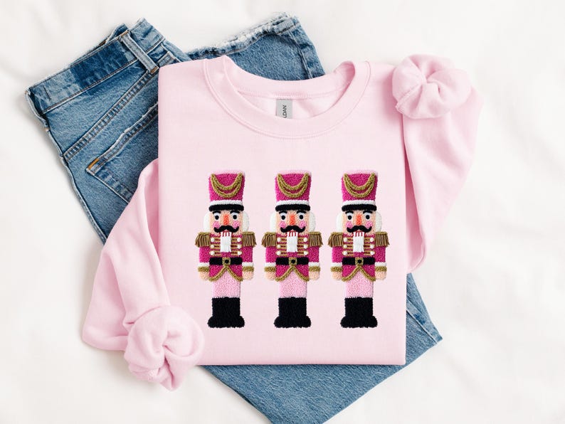 May include: A light pink sweatshirt featuring three embroidered nutcracker figures. The nutcrackers have pink and gold uniforms, black boots, and detailed facial features. The sweatshirt is laid on top of a pair of blue jeans.