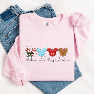 May include: A light pink sweatshirt with a festive design featuring four Mickey Mouse head shapes. The designs include candy canes, snowflakes, and a gingerbread cookie. The text "Mickey's Very Merry Christmas" is written below the designs.