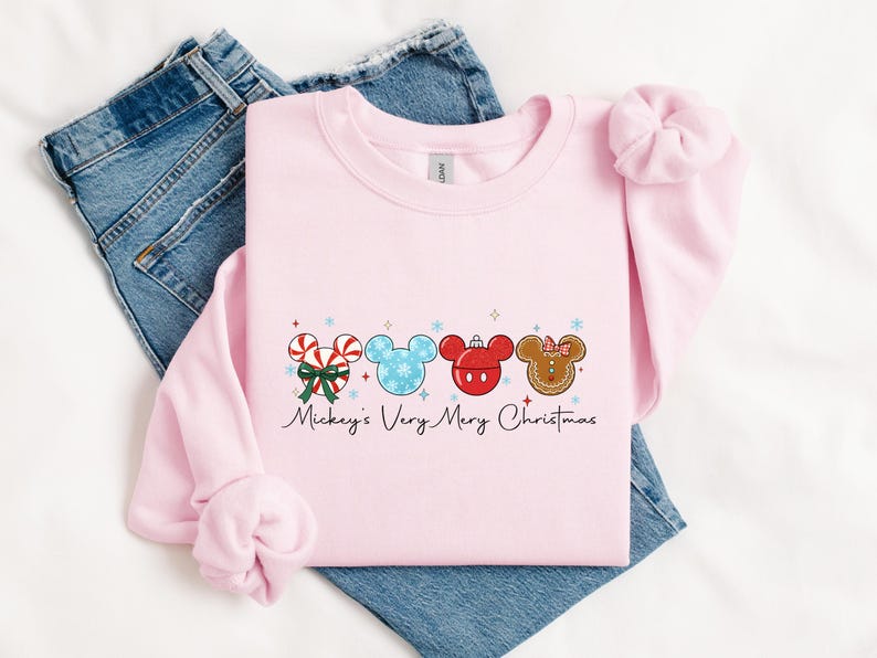 May include: A light pink sweatshirt with a festive design featuring four Mickey Mouse head shapes. The designs include candy canes, snowflakes, and a gingerbread cookie. The text "Mickey's Very Merry Christmas" is written below the designs.