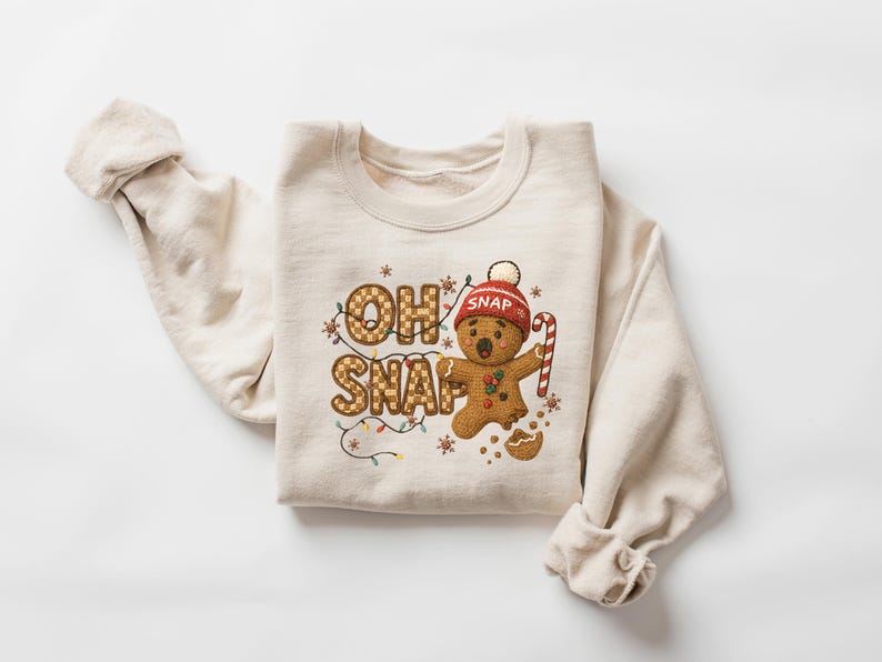 K&ouml;nnte beinhalten: Beiges Sweatshirt mit dem Schriftzug "OH SNAP" in Lebkuchen-Schrift. Ein Lebkuchenmann mit roter M&uuml;tze und Zuckerstange ist abgebildet. Das Design umfasst Lichterketten und Sterne, ideal f&uuml;r die Weihnachtszeit.