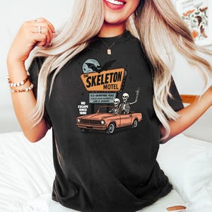 May include: Black t-shirt featuring a vintage-style illustration of a motel sign that reads "The Skeleton Motel." The design includes a car with skeletons and the text "Old Graveyard Road." The shirt also has the text "No Escape Since 1690."