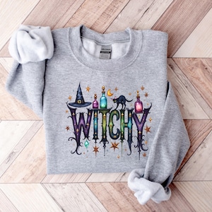May include: A light gray sweatshirt with a colorful graphic that says "WITCHY". The graphic includes a witch's hat, potion bottles, a black cat, and stars. The sweatshirt is made of a soft material.