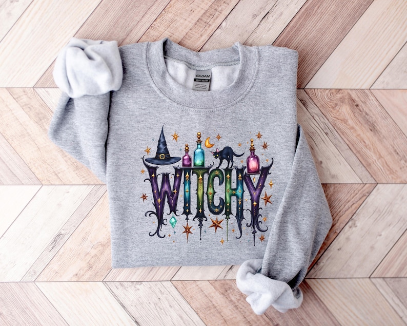 May include: A light gray sweatshirt with a colorful graphic that says "WITCHY". The graphic includes a witch's hat, potion bottles, a black cat, and stars. The sweatshirt is made of a soft material.