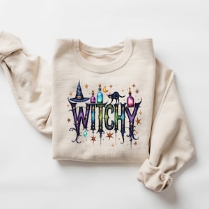 May include: A cream-colored sweatshirt with the word "WITCHY" in a colorful, whimsical design. The design includes a witch hat, potion bottles, a black cat, and stars, perfect for Halloween or a witchy aesthetic.