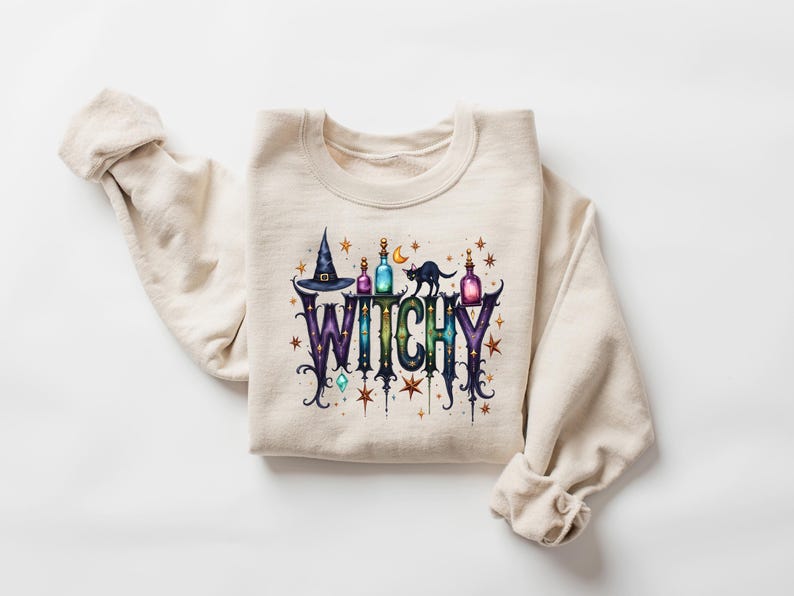 May include: A cream-colored sweatshirt with the word "WITCHY" in a colorful, whimsical design. The design includes a witch hat, potion bottles, a black cat, and stars, perfect for Halloween or a witchy aesthetic.