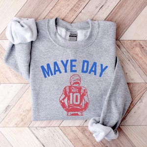 K&ouml;nnte beinhalten: Ein hellgraues Sweatshirt mit dem blauen Text "MAYE DAY". Darunter befindet sich eine rote Illustration eines Footballspielers mit der Nummer 10. Das Sweatshirt liegt auf einer Holzoberfl&auml;che.