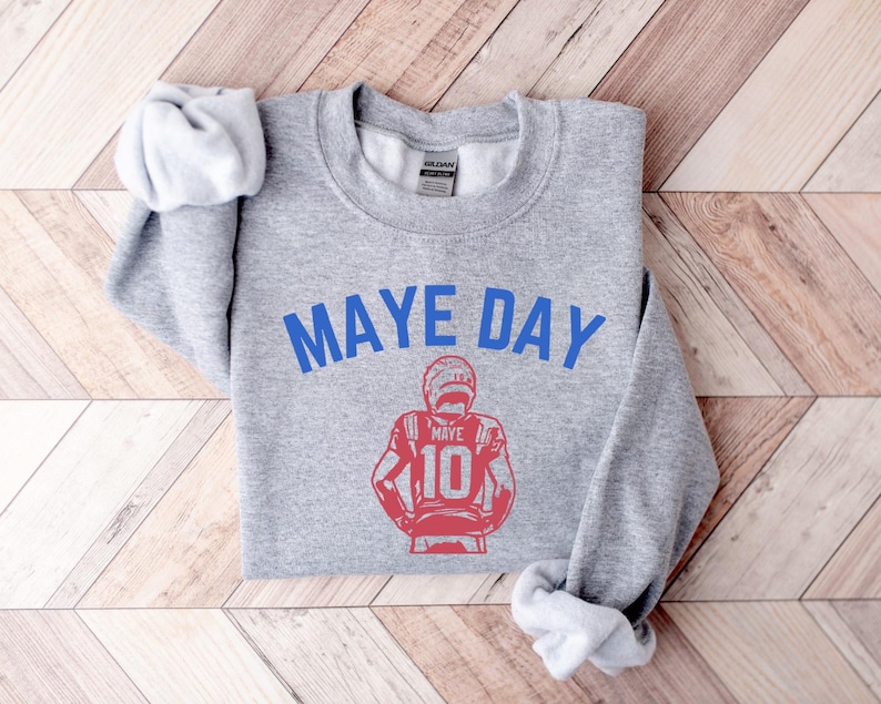 K&ouml;nnte beinhalten: Ein hellgraues Sweatshirt mit dem blauen Text "MAYE DAY". Darunter befindet sich eine rote Illustration eines Footballspielers mit der Nummer 10. Das Sweatshirt liegt auf einer Holzoberfl&auml;che.