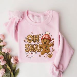 K&ouml;nnte beinhalten: Ein hellrosa Sweatshirt mit dem Schriftzug "OH SNAP" in einem Karomuster, das einen Lebkuchenmann mit roter M&uuml;tze und Zuckerstange zeigt. Das Design umfasst Lichterketten und einen zerbrochenen Keks.