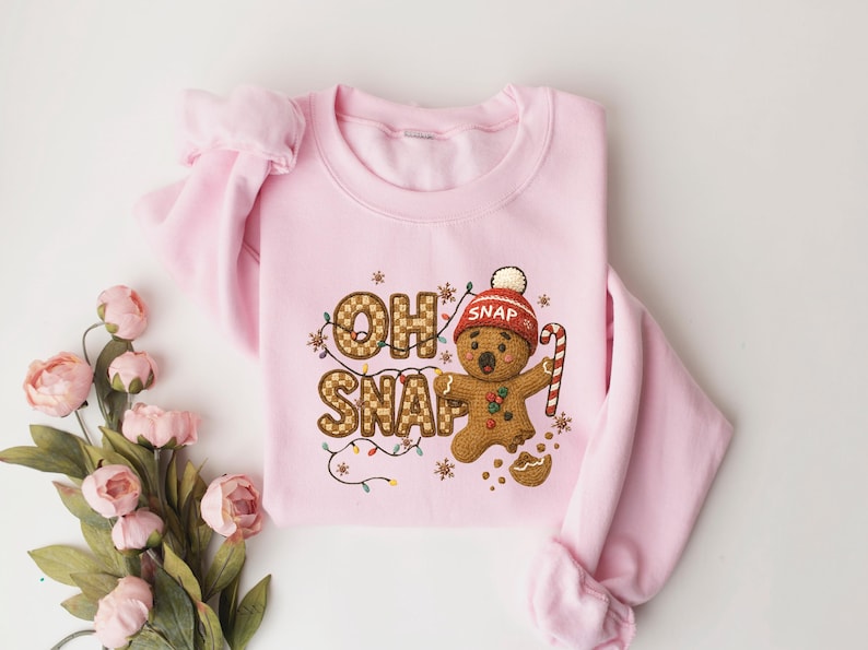 K&ouml;nnte beinhalten: Ein hellrosa Sweatshirt mit dem Schriftzug "OH SNAP" in einem Karomuster, das einen Lebkuchenmann mit roter M&uuml;tze und Zuckerstange zeigt. Das Design umfasst Lichterketten und einen zerbrochenen Keks.