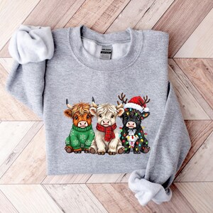 May include: A grey sweatshirt with a graphic of three Highland cows. One cow is in a green sweater, another in a red scarf, and the third in a Santa hat and Christmas lights. The cows are brown, white, and black.