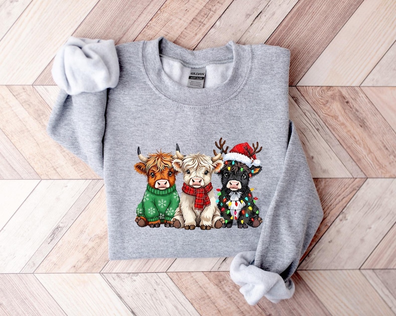 May include: A grey sweatshirt with a graphic of three Highland cows. One cow is in a green sweater, another in a red scarf, and the third in a Santa hat and Christmas lights. The cows are brown, white, and black.