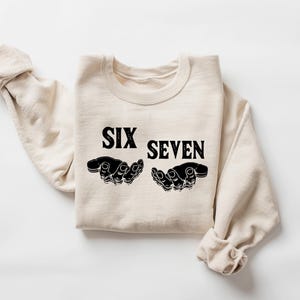 May include: A cream-colored sweatshirt with the words "SIX SEVEN" in bold black letters. Below the text are two black hand illustrations, palms up. The sleeves are rolled up, and the sweatshirt is laid flat on a white surface.