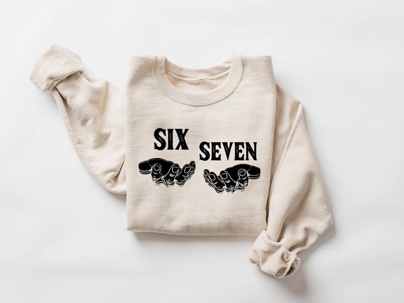 May include: A cream-colored sweatshirt with the words "SIX SEVEN" in bold black letters. Below the text are two black hand illustrations, palms up. The sleeves are rolled up, and the sweatshirt is laid flat on a white surface.