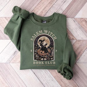 May include: Green sweatshirt with the text "Salem Witch Book Club" and an illustration of a witch reading on a pumpkin in front of a full moon. The design is in a vintage style, with the words "All Witches Welcome" at the bottom.
