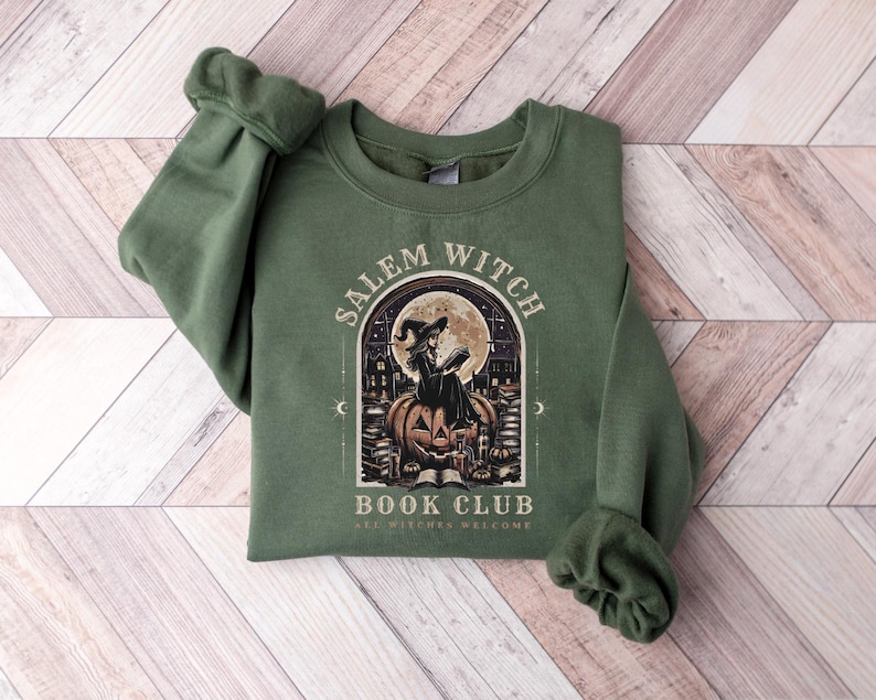 May include: Green sweatshirt with the text "Salem Witch Book Club" and an illustration of a witch reading on a pumpkin in front of a full moon. The design is in a vintage style, with the words "All Witches Welcome" at the bottom.