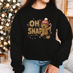 K&ouml;nnte beinhalten: Schwarzes Sweatshirt mit einem Lebkuchenmann-Design. Der Text "OH SNAP" ist in einem Karomuster gehalten, mit einem Lebkuchenmann, der eine M&uuml;tze tr&auml;gt und eine Zuckerstange h&auml;lt. Ideal f&uuml;r die Weihnachtszeit.