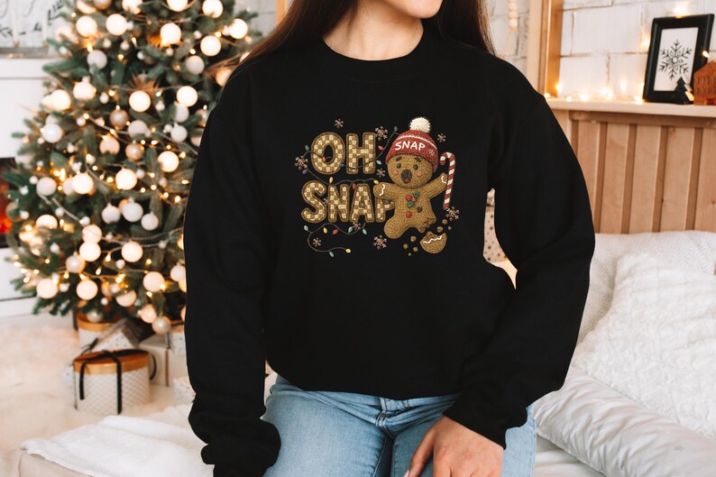 K&ouml;nnte beinhalten: Schwarzes Sweatshirt mit einem Lebkuchenmann-Design. Der Text "OH SNAP" ist in einem Karomuster gehalten, mit einem Lebkuchenmann, der eine M&uuml;tze tr&auml;gt und eine Zuckerstange h&auml;lt. Ideal f&uuml;r die Weihnachtszeit.