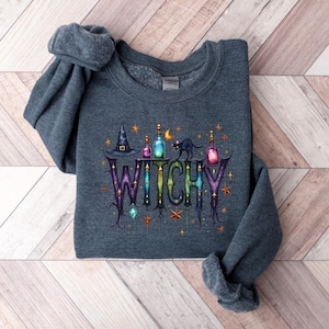 May include: A dark gray sweatshirt with the word "WITCHY" in a decorative font. The design includes a witch's hat, potion bottles, a black cat, and stars. The sweatshirt is folded on a light wood-grain surface.
