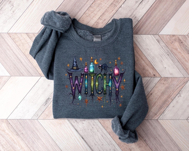 May include: A dark gray sweatshirt with the word "WITCHY" in a decorative font. The design includes a witch's hat, potion bottles, a black cat, and stars. The sweatshirt is folded on a light wood-grain surface.
