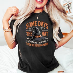 May include: Black t-shirt with the text "SOME DAYS YOU JUST HAVE TO PUT ON THE HAT AND REMIND THEM WHO THEY'RE DEALING WITH". The graphic features a witch's hat with a buckle and spiderwebs. A Halloween-themed design.