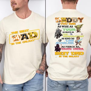 Puede incluir: Camiseta color crema con el texto "THE BEST DAD IN THE GALAXY" en la parte delantera. La parte trasera presenta "DADDY" y frases que comparan al destinatario con Yoda, The Mandalorian, Luke Skywalker y Darth Vader.