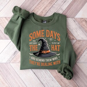 May include: Green sweatshirt with the text "SOME DAYS you just have the HAT and remind them who they're dealing with." The design includes a black witch hat with an orange buckle, bats, and stars. The sweatshirt is folded on a wooden surface.