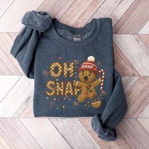 K&ouml;nnte beinhalten: Ein marineblaues Sweatshirt mit dem gestickten Schriftzug "OH SNAP" und einem Lebkuchenmann mit roter M&uuml;tze und Zuckerstange. Das Design umfasst dekorative Lichter und Buchstaben im Lebkuchen-Stil.
