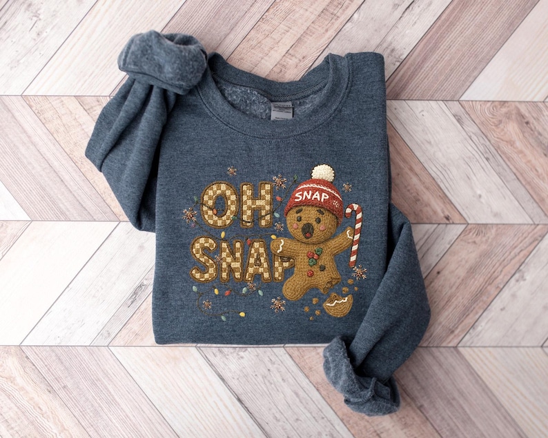 K&ouml;nnte beinhalten: Ein marineblaues Sweatshirt mit dem gestickten Schriftzug "OH SNAP" und einem Lebkuchenmann mit roter M&uuml;tze und Zuckerstange. Das Design umfasst dekorative Lichter und Buchstaben im Lebkuchen-Stil.