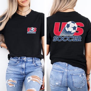 Puede incluir: Una camiseta negra con un dise&ntilde;o gr&aacute;fico que incluye el texto US SOCCER. La parte delantera muestra un peque&ntilde;o logotipo en el pecho izquierdo, mientras que la parte trasera presenta una versi&oacute;n grande del mismo dise&ntilde;o con un bal&oacute;n y letras rojas y azules.