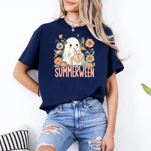 Op de afbeelding: Marineblauwe t-shirt met een "Summerween" ontwerp. De afbeelding toont een spook dat een drankje drinkt, omringd door bloemen. De tekst "Summerween" is oranje. Het t-shirt wordt gedragen met een blauwe jeans.