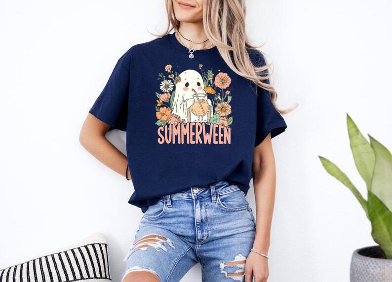 Op de afbeelding: Marineblauwe t-shirt met een "Summerween" ontwerp. De afbeelding toont een spook dat een drankje drinkt, omringd door bloemen. De tekst "Summerween" is oranje. Het t-shirt wordt gedragen met een blauwe jeans.