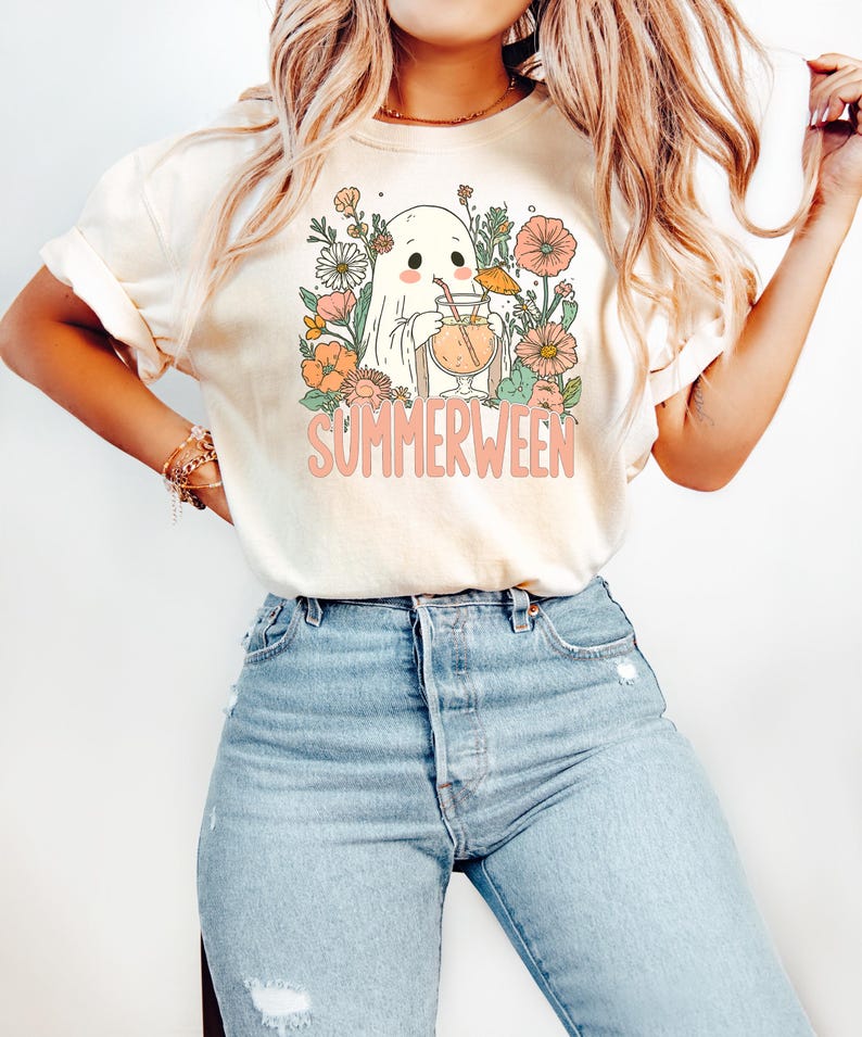 Op de afbeelding: Cr&egrave;mekleurig T-shirt met een cartoongeest die een cocktail vasthoudt, omringd door bloemen. De roze tekst luidt "SUMMERWEEN". Het shirt wordt gecombineerd met lichtblauwe jeans.