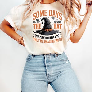 May include: Cream-colored t-shirt with the text "Some Days You Just Have To Put On The Hat And Remind Them Who They're Dealing With." The graphic features a witch's hat, spider, and bats.