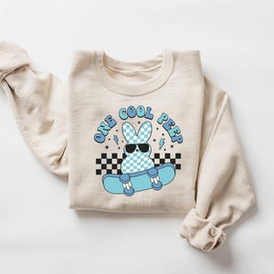 May include: A cream-coloured sweatshirt featuring a graphic of a bunny wearing sunglasses and riding a skateboard. The text "ONE COOL PEEP" is above the bunny in blue. The bunny has a blue and white chequered pattern.