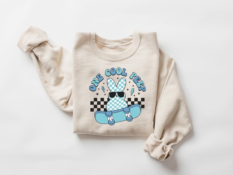 May include: A cream-coloured sweatshirt featuring a graphic of a bunny wearing sunglasses and riding a skateboard. The text "ONE COOL PEEP" is above the bunny in blue. The bunny has a blue and white chequered pattern.