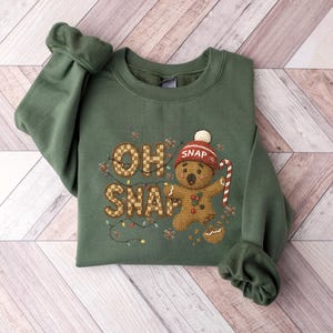 K&ouml;nnte beinhalten: Gr&uuml;nes Sweatshirt mit dem Schriftzug "OH SNAP" und einem Lebkuchenmann-Design. Der Lebkuchenmann tr&auml;gt einen roten Hut und h&auml;lt eine Zuckerstange. Das Design umfasst festliche Lichter und Lebkuchenkr&uuml;mel.