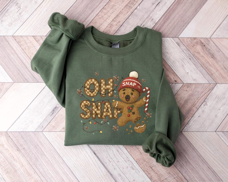 K&ouml;nnte beinhalten: Gr&uuml;nes Sweatshirt mit dem Schriftzug "OH SNAP" und einem Lebkuchenmann-Design. Der Lebkuchenmann tr&auml;gt einen roten Hut und h&auml;lt eine Zuckerstange. Das Design umfasst festliche Lichter und Lebkuchenkr&uuml;mel.