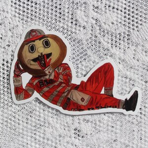 May include: A sticker featuring a cartoon mascot in a red and gray striped uniform and hat, reclining on a white surface. The mascot has a brown head and is wearing black shoes. The word "Ohio" is visible on the uniform.