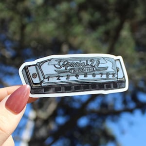 May include: A sticker of a harmonica with the text "Special 20 Progressive" and numbers 1-9. The harmonica is light blue and gray with black accents. The sticker is held up against a blurred green and blue background.