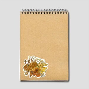 May include: A spiral-bound notepad with a brown cover. A colorful sticker of a lionfish is affixed to the bottom left corner. The lionfish has orange, yellow, and white details. The notepad is blank and ready for notes.